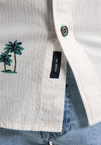 Camicia bianca con tessuto testurizzato, caratterizzata da palme ricamate in verde e marrone, e un'etichetta scura con la scritta "PETROL NO." sul lato.