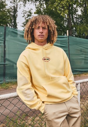 Yellow hoodie with front pocket, drawstring, and logo. Features textured patches on sleeves. Paired with beige trousers.