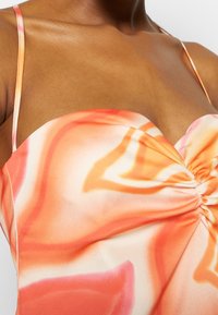 Gros plan d'une personne portant une robe sans manches avec de fines bretelles et un motif tourbillonnant abstrait orange, blanc et rose.