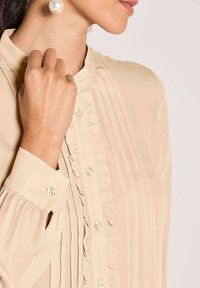 Beige Bluse mit vertikalen Streifen, Rüschendetails entlang der Knopfleiste und Perlenknöpfen. Glatter, leichter Stoff; enganliegende Manschetten.