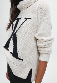 Jasna, wełniana dzianinowa sweter z czarnym logo "K" z przodu. Posiada golf i prążkowany dół z bocznymi rozcięciami. Miękka tekstura.