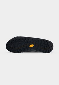Semelle noire robuste de chaussure avec des rainures profondes et un logo hexagonal jaune au centre, présentée sur un fond clair uni.