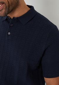 Námořnická modrá textured polo košile s krátkými rukávy, se vzorem z pletené tkaniny a třemi černými knoflíky na límci.