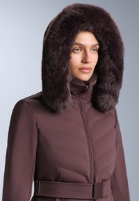 Žena u mauve podstavljenom zimskom kaputu s velikim tamnim kapuljačom obloženim krznenim rubom i remenom u struku, na jednostavnoj sivoj pozadini.