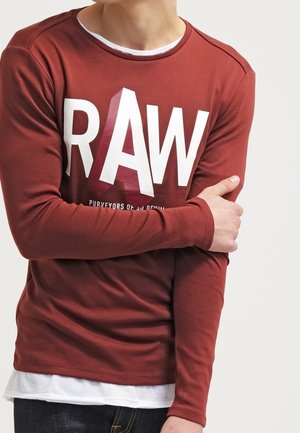 Homme portant une chemise à manches longues couleur rouille avec un grand texte blanc "RAW", superposée à un sous-vêtement blanc, tenant son bras gauche.