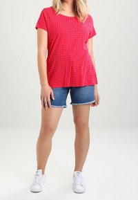 Top corto rojo de manga corta con lunares blancos, combinado con pantalones cortos de mezclilla. Zapatillas blancas completan el conjunto casual. Tela suave, forma ajustada.