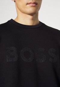 Man som bär en svart tröja med rund halsringning där "BOSS" är stavat med små svarta nitar över bröstet.