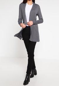 Harmaaraidallinen neuletakki avoimella etuosalla, pitkät hihat ja pehmeä koostumus, yhdistetty mustiin istuviin housuihin ja nilkkureihin.