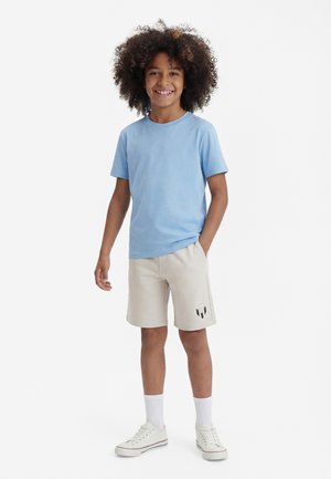 Smilende barn med krøllete hår iført lyseblå t-skjorte, beige shorts med svart logo, hvite sokker og hvite joggesko, stående med den ene hånden i lomma.