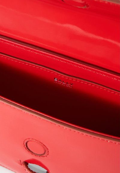 Inuti en röd lackläderväska med en slät yta, där en synlig varumärkeslogo är präglad på en läderetikett och en cirkulär utskärning är synlig.