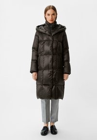 Donkergroene gewatteerde jas met een capuchon, met een quilted ontwerp, lange mouwen en een ritssluiting aan de voorkant, gedragen over een grijze broek en zwarte schoenen.