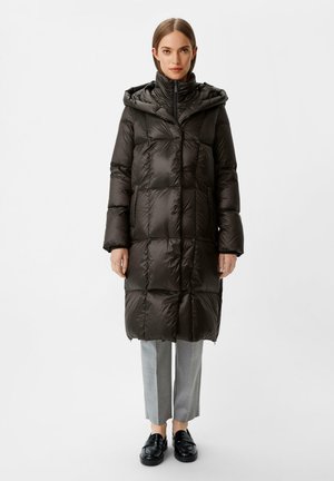 Donkergroene gewatteerde jas met een capuchon, met een quilted ontwerp, lange mouwen en een ritssluiting aan de voorkant, gedragen over een grijze broek en zwarte schoenen.