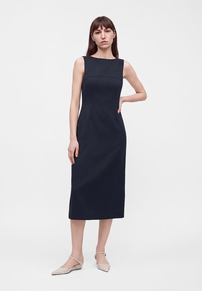 Femme debout portant une robe midi noire sans manches et des chaussures plates beiges sur un fond clair uni.