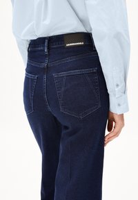 Vaqueros de mezclilla azul oscuro con una cintura alta, diseño de cinco bolsillos y una etiqueta negra en la parte trasera. Textura suave y corte recto.