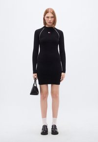 Μαύρο μακρυμάνικο φόρεμα με ριμπ ύφανση, λευκές λεπτομέρειες και μικρό λογότυπο· συνδυασμένο με μαύρη τσάντα χειρός και μαύρα chunky παπούτσια, με λευκές κάλτσες.