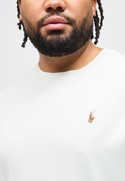 Άνδρας με γενειάδα και πλεγμένα μαλλιά, φορώντας λευκό μπλουζάκι με στρογγυλή λαιμόκοψη που φέρει ένα μικρό κεντητό λογότυπο παίκτη πολο στο στήθος.