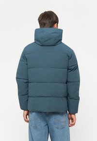 Teal puffer jacket with a hood, featuring a quilted design and discreet stitching. Paired with light blue jeans.