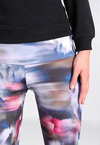 Leggings con estampado en una mezcla de colores que incluyen azul, rosa y gris, fabricados con un tejido suave y elástico; combinados con un sudadera negra.