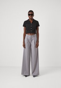 Chemise noire cintrée à manches courtes avec des boutons, assortie à un pantalon large plissé gris et des lunettes de soleil sombres. Texture lisse sur toute la surface.