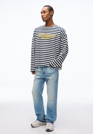 Jonge man die een zwart-wit gestreept shirt met lange mouwen en gele tekst draagt, en een lichtblauwe spijkerbroek, staand met één hand in de zak.