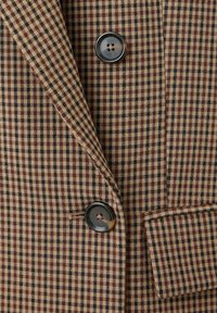 Veste à carreaux marron et noire avec deux boutons noirs et une poche à rabat en bas à droite.
