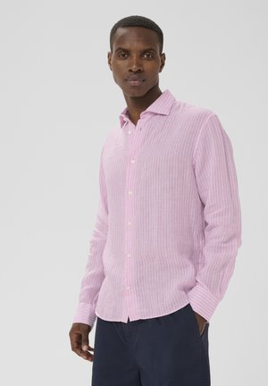 Homme portant une chemise à manches longues rayée rose et blanche, boutonnée, avec un pantalon bleu foncé, debout devant un fond clair uni.