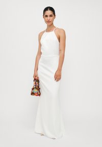 White halter neck dress with a fitted silhouette and a slight train, held with a colorful beaded bag. Smooth texture, minimal detailing.