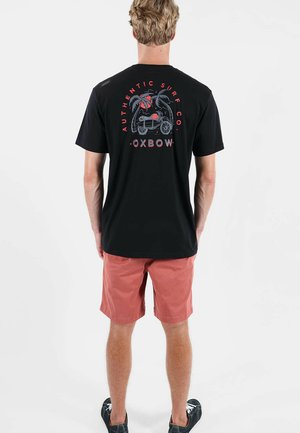 Homme portant un t-shirt noir avec un logo et un texte sur le thème du surf en rouge et gris, associé à un short corail et des baskets noires, debout devant un fond blanc.