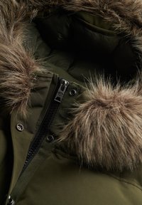 Gros plan sur un manteau d'hiver vert olive avec une fermeture éclair et une bordure en fausse fourrure sur la capuche, montrant des détails du tissu et des coutures.