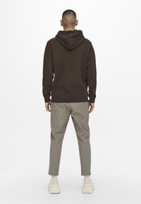 Hombre de pie de espaldas, vistiendo una sudadera oscura marrón, pantalones claros grises, calcetines blancos y zapatillas gruesas beige, sobre un fondo blanco.