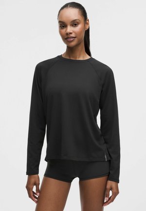 Femme debout portant un haut à manches longues noir et un short noir sur un fond clair uni, regardant devant avec un léger sourire.