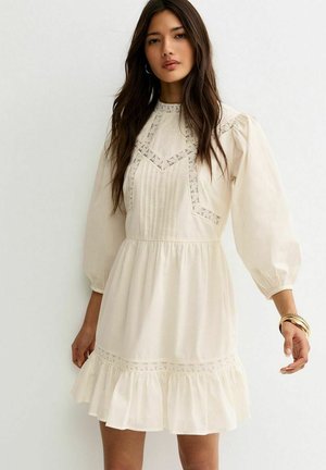 Kobieta o długich ciemnych włosach, ubrana w kościelnobiałą sukienkę z długim rękawem, z koronkowymi detalami, plisami na gorsecie i falbankami na dole, stojąca na białym tle.