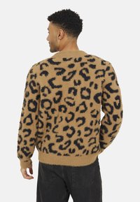 Pull en beige peluche avec imprimé léopard noir, poignets et ourlet côtelés, texture semblable à de la laine, silhouette ajustée. Porté avec un pantalon sombre.