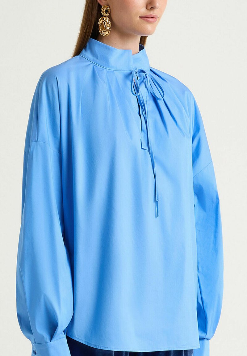 Blouse bleu clair avec un col montant, des manches longues et un détail froncé au niveau du cou. Entièrement en tissu lisse, avec une fermeture à nœud papillon.