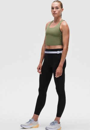 Haut de sport coupé vert olive associé à des leggings noirs taille haute. Le modèle porte des chaussures de sport gris clair avec des semelles jaunes.