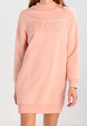 Femme portant une robe sweat-shirt rose clair à manches longues avec "VOLCOM" brodé sur la poitrine.