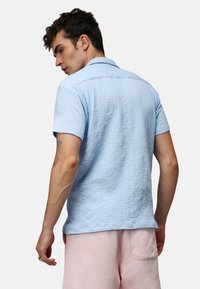Camisa de manga corta azul claro con una tela texturizada, cuello puntiagudo y dobladillo recto, combinada con unos pantalones cortos rosa claro.