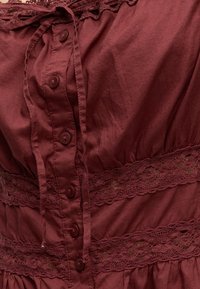 Tissu bordeaux avec boutons au centre, comprenant des bandes de dentelle et un cordon noué sur le bord supérieur.