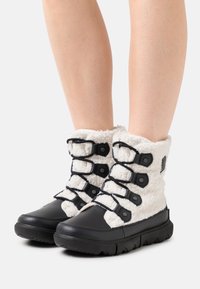 Botas difusas en blanco y negro con múltiples correas negras, exhibidas sobre piernas desnudas contra un fondo blanco liso.