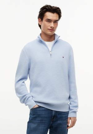 Ung mann iført lys blå teksturert genser med kvartglidelås over hvit skjorte og blå jeans, stående med den ene hånden i lommen.