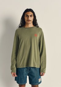 Langærmet olivengrøn bomulds skjorte med et rødt logo på venstre bryst, parret med denimshorts med et mærke på siden.