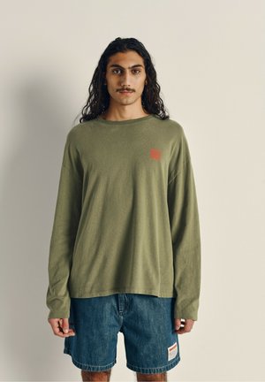 Langärmliges olivgrünes Baumwollhemd mit rotem Logo auf der linken Brust, kombiniert mit Jeansshorts, die ein Etikett an der Seite haben.