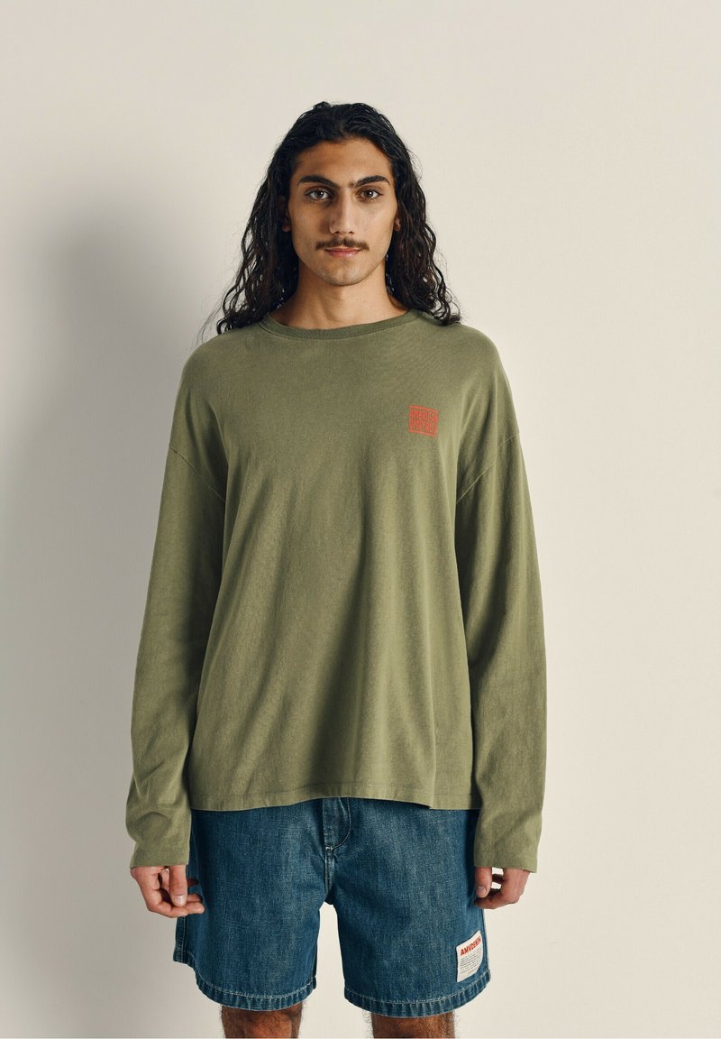 Langærmet olivengrøn bomulds skjorte med et rødt logo på venstre bryst, parret med denimshorts med et mærke på siden.
