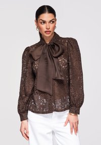 Femme aux cheveux tirés en arrière portant un chemisier en dentelle florale marron transparent avec un grand nœud en satin, des manches bouffantes, et un pantalon blanc, posant devant un fond gris.