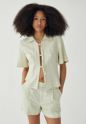 Vrouw met grote krullende haren draagt een lichtgroen geruit T-shirt met korte mouwen en bijpassende korte broek, met handen in de zakken tegen een effen achtergrond.