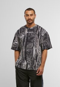 Mand med kort hår og øreringe iført en oversized sort-hvid mønstret skjorte med "Colucci" logo og sorte bukser, stående foran en hvid baggrund.