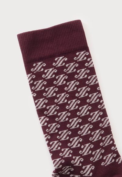 Chaussette marron foncé avec motif stylisé blanc répété "H" et large bord-côte sur un fond clair uni.