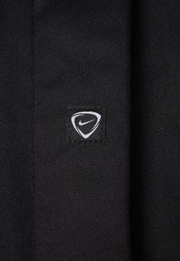 Zwart gestructureerde stof met een klein vierkant Nike-logo label waarop een witte swoosh staat, omrand door een gebogen driehoek.