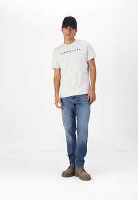 Graues Baumwoll-T-Shirt mit "TOMMY JEANS"-Logo; blaue Jeans; braune Wildlederschuhe; schwarze Mütze. Modell steht vor einem schlichten weißen Hintergrund.