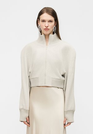 Žena oblečená v béžovom svetri na zips s nadýchanými ramenami a dlhými manžetami, ktoré kombinuje s ľahkou béžovou saténovou sukňou a visiace krištáľové náušnice.